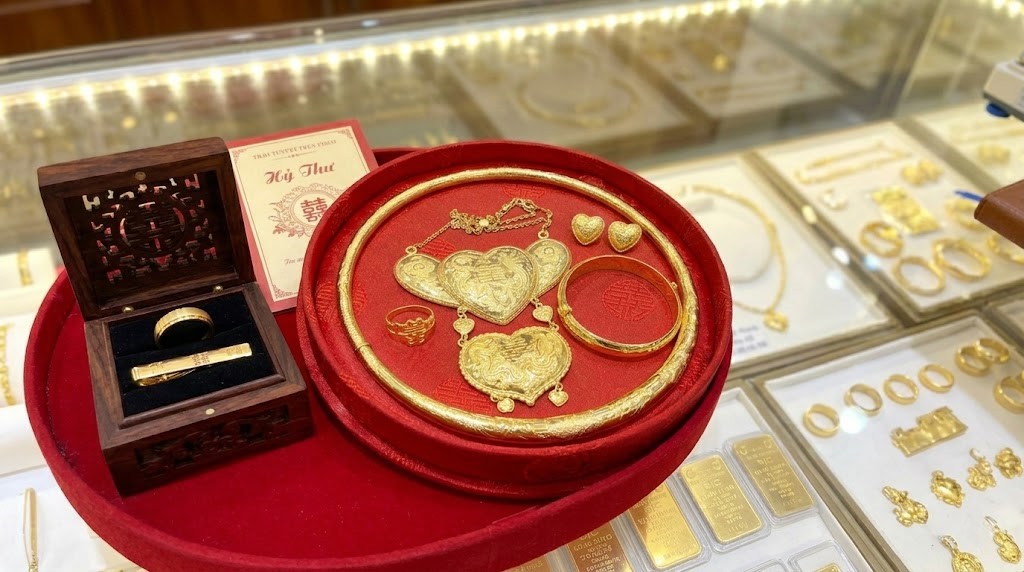 Trưng bày bộ trang sức cưới vàng 24K truyền thống Việt Nam trên khay nhung đỏ. Bộ sản phẩm bao gồm kiềng cổ, dây chuyền mặt trái tim, bông tai, nhẫn và một hộp gỗ mở chứa nhẫn nam và kẹp cà vạt vàng